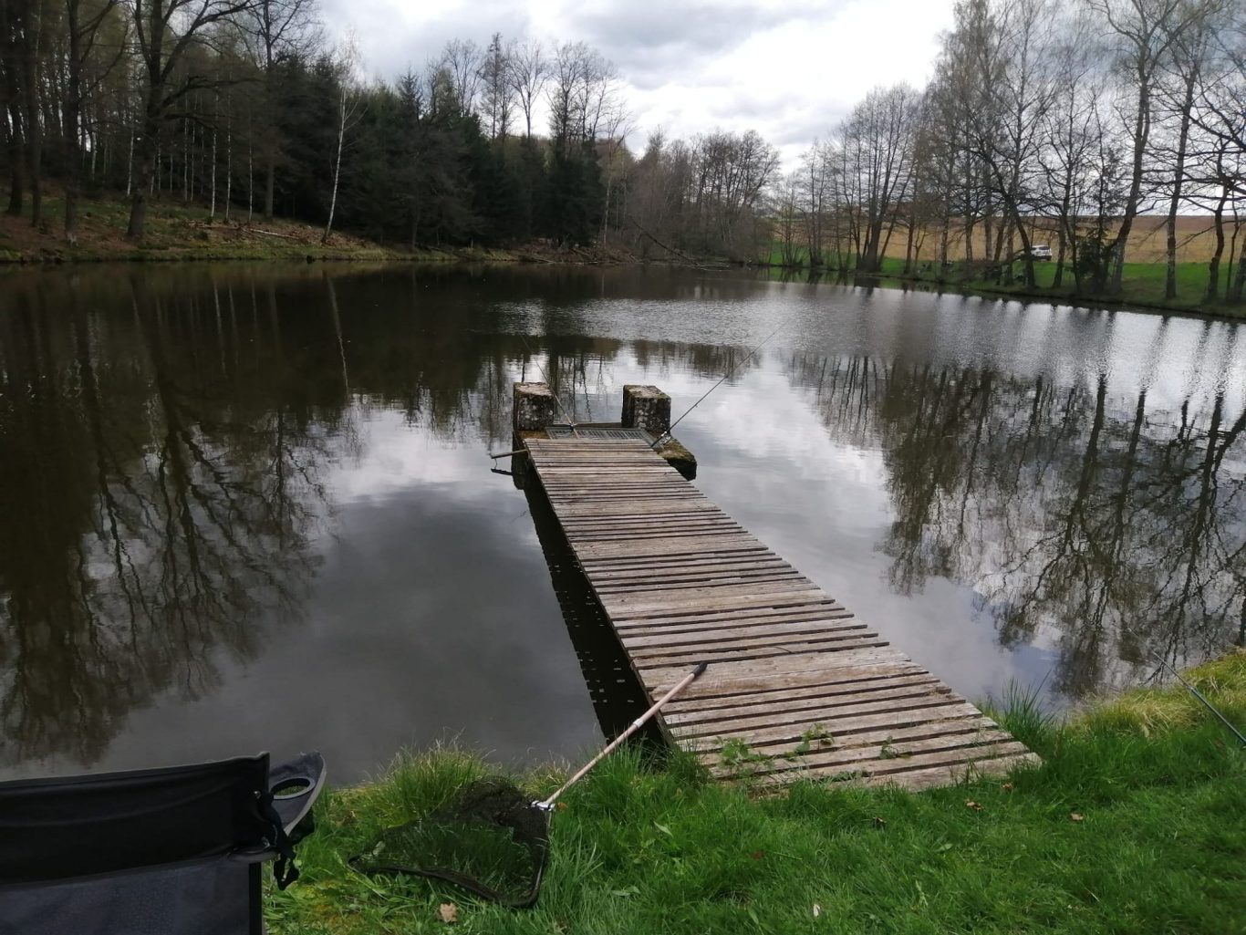 Ein Holzsteg führt zu einem ruhigen Teich, umgeben von Bäumen und Wiese. Ein Holzsteg führt zu einem ruhigen Teich, umgeben von Bäumen und Wiese.