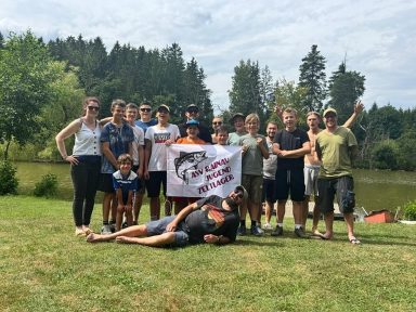 Gruppe junger Menschen posiert am Ufer eines Sees mit einem Banner.