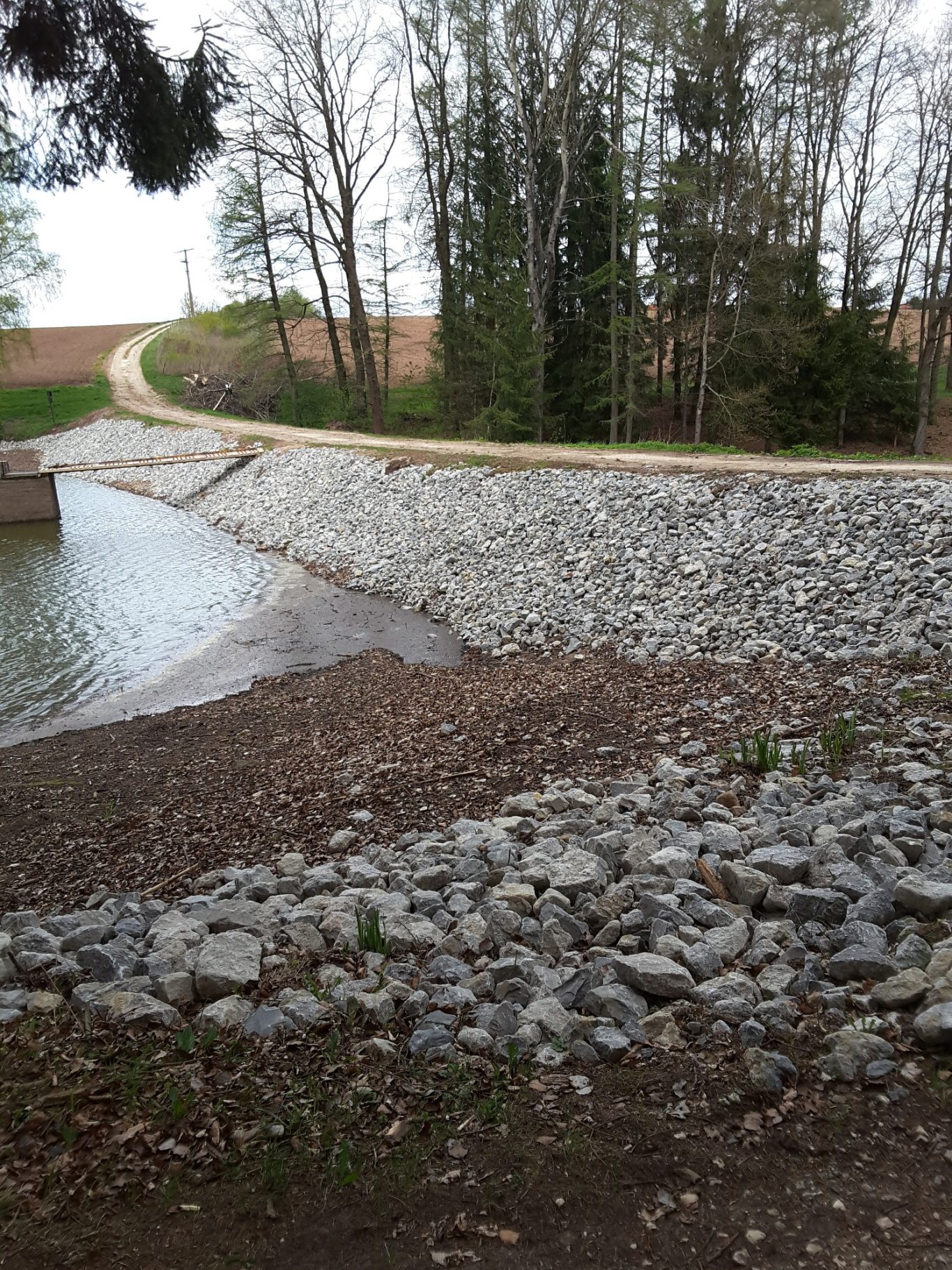 Reparaturen an Staumauern und Wehren Uferbereich mit Kies, schmaler Weg und Bäume im Hintergrund.