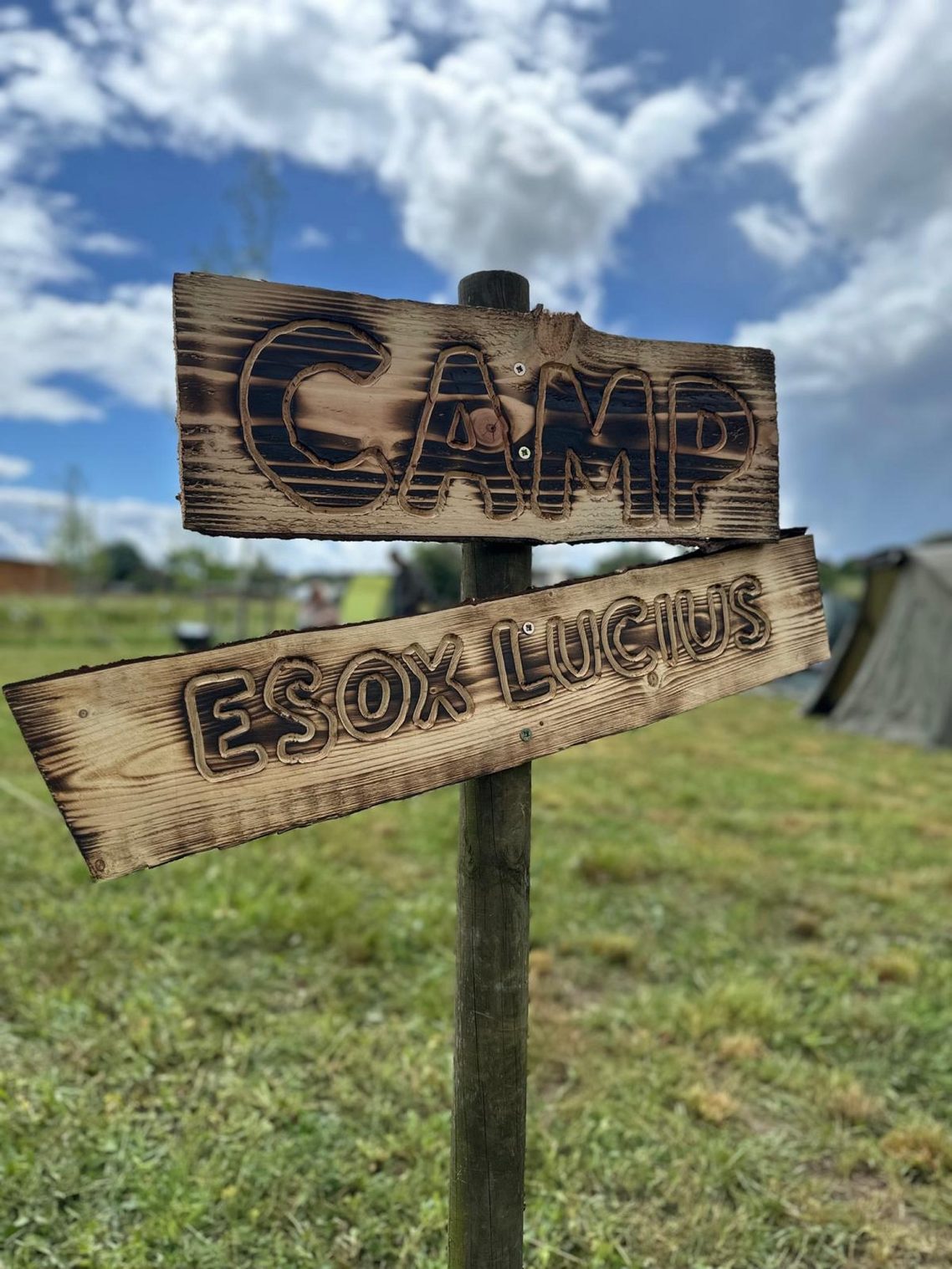 Camp Esox Lucius (Hecht) Holzschild mit der Aufschrift "CAMP" und "EINRICHTUNG" im Freien.