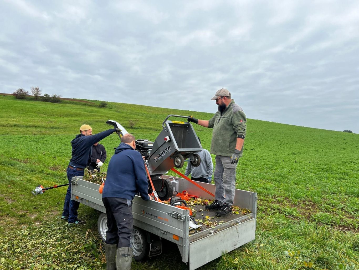 Drei Personen arbeiten auf einem Feld und bedienen Maschinen auf einem Anhänger.