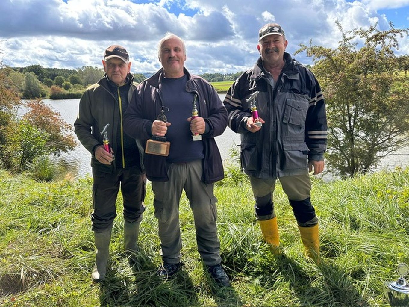 Drei Männer stehen am Ufer eines Gewässers, jeweils mit Angelgeräten in der Hand.