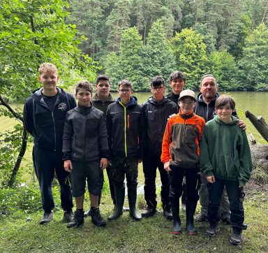 Gruppe von zehn Personen am Ufer eines Sees in einem Waldgebiet. Alle tragen Freizeitkleidung.
