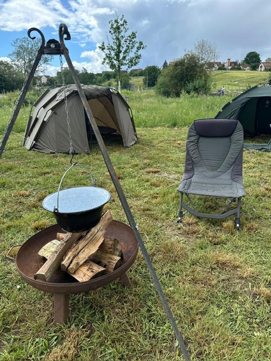 Auch beim Angeln kann man es gemütlich haben. Feuerstelle mit Holzscheiten, Campingstühle und Zelte in einer grünen Wiese.