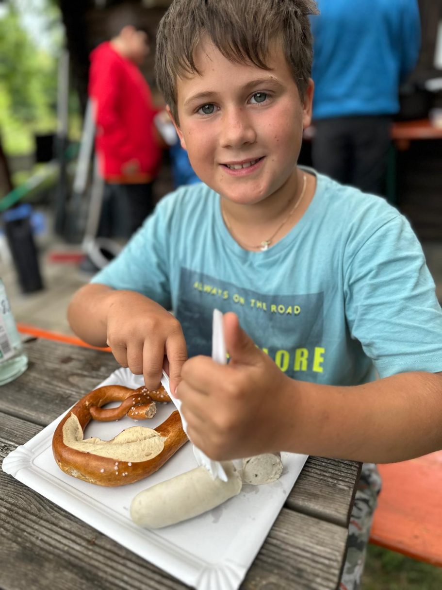 Gemeinsam feiern Junge mit Brezel und Würstchen, lächelt und zeigt Daumen hoch.