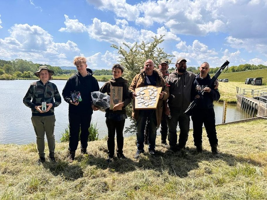 Wettkampf Gruppe von acht Personen steht am Ufer eines Gewässers, umgeben von Bäumen und Wolken.